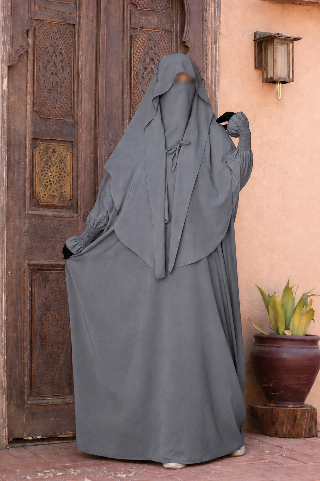 Jilbab 2 pièces gris en crêpe royal avec khimar long et jupe ample, porté de face par un mannequin devant une porte orientale en bois.