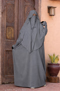 Jilbab 2 pièces gris en crêpe royal avec khimar long et jupe ample, porté de face par un mannequin devant une porte orientale en bois.