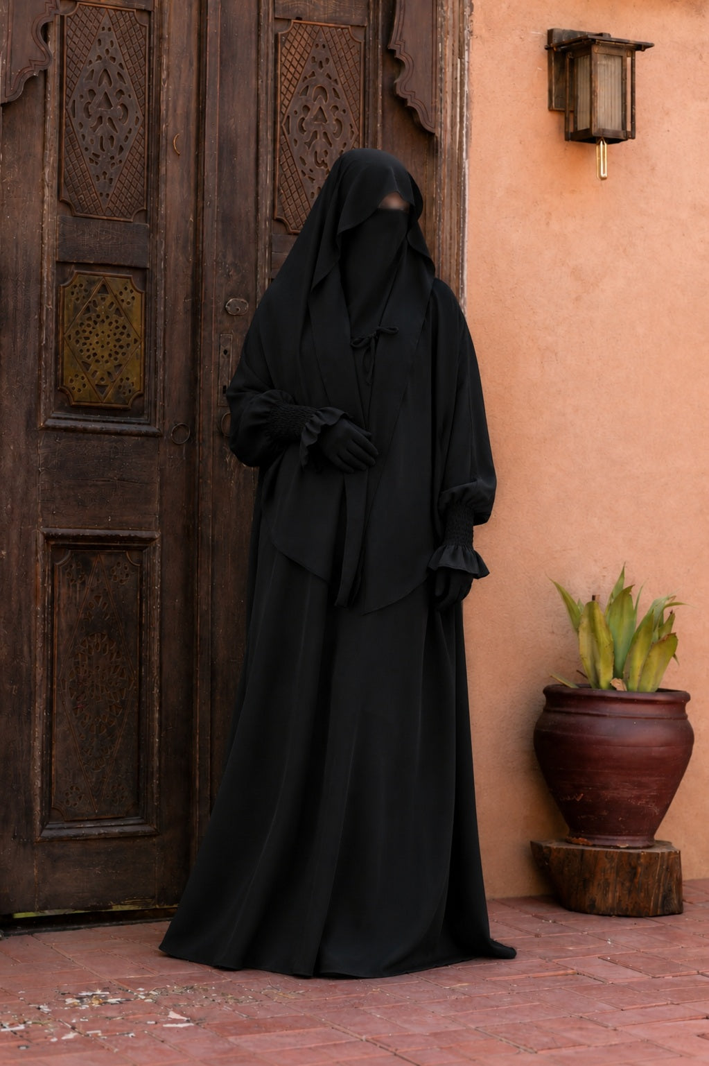 Jilbab 2 pièces noir en crêpe royal avec khimar long couvrant et jupe ample, porté de face devant une porte orientale en bois sculpté.
