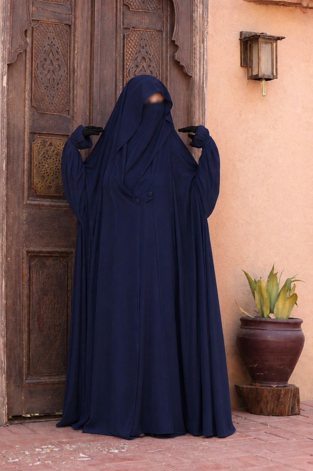 Jilbab Liyane bleu nuit avec deux boutons et manches resserrées en crêpe royale, porté de face devant une porte orientale.