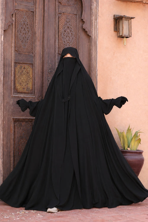 Jilbab Liyane noir à deux boutons en crêpe royale, femme présentant l'ampleur et la fluidité du tissu avec une coupe mastour élégante, manches élastiquées et taille unique adaptée aux personnes mesurant entre 1m55 et 1m75.