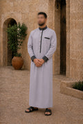 Homme portant un qamis gris clair premium avec col officier gris anthracite, coupe longue élégante et détails raffinés dans un décor d'inspiration méditerranéenne.