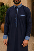 Qamis premium homme bleu marine avec col, poignets et patte de boutonnage bleu ciel contrastés, boutons carrés dorés et poche poitrine, présenté dans un décor méditerranéen.